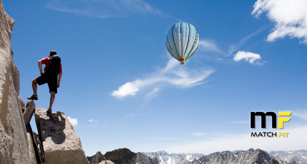 person climbing sheer rockface on left, looking out at hot air balloon floating in blue sky with wispy white clouds